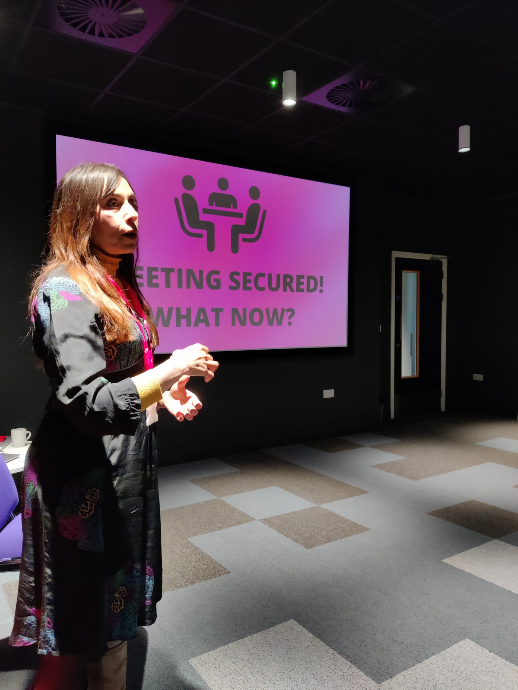 Flapjack festival - Woman stands up in front of a screen with a pink background, with a small diagram showing seated people, and text which reads: "Meeting Secured What Now?"